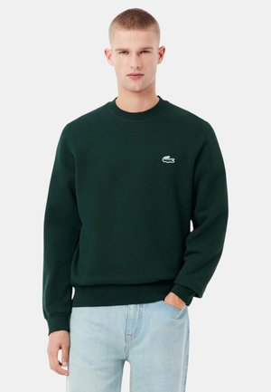 Felpa verde scuro con scollo rotondo, polsini e orlo a costine. Presenta un piccolo logo Lacoste bianco sul petto a sinistra. Tessuto in misto cotone.