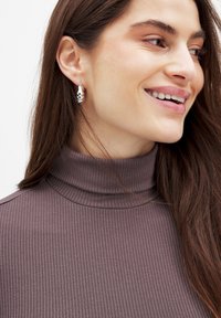 Silver hoops med en skulptural design, bärs av en person med långt mörkt hår, i en tight, ribbad, brun polotröja.