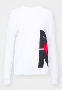 Pull à manches longues blanc avec un motif abstrait audacieux en noir et rouge sur le côté droit, comprenant une forme de cœur dans le motif.