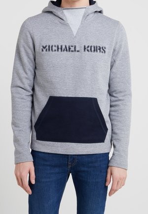 Man wearing grey waffle-knit hoodie with navy kangaroo pocket and "MICHAEL KORS" logo, paired with blue jeans, against white background.