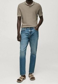 Mann in beigem, kurzärmeligem Strickpolo, blauen Jeans und beigen Sandalen, der mit einer Hand in der Tasche vor einem hellen Hintergrund steht.