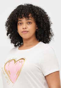 Tricou alb din bumbac, cu un grafic mare în formă de inimă roz conturat în aur, având textul "minte calmă, inimă puternică" într-un font jucăuș.