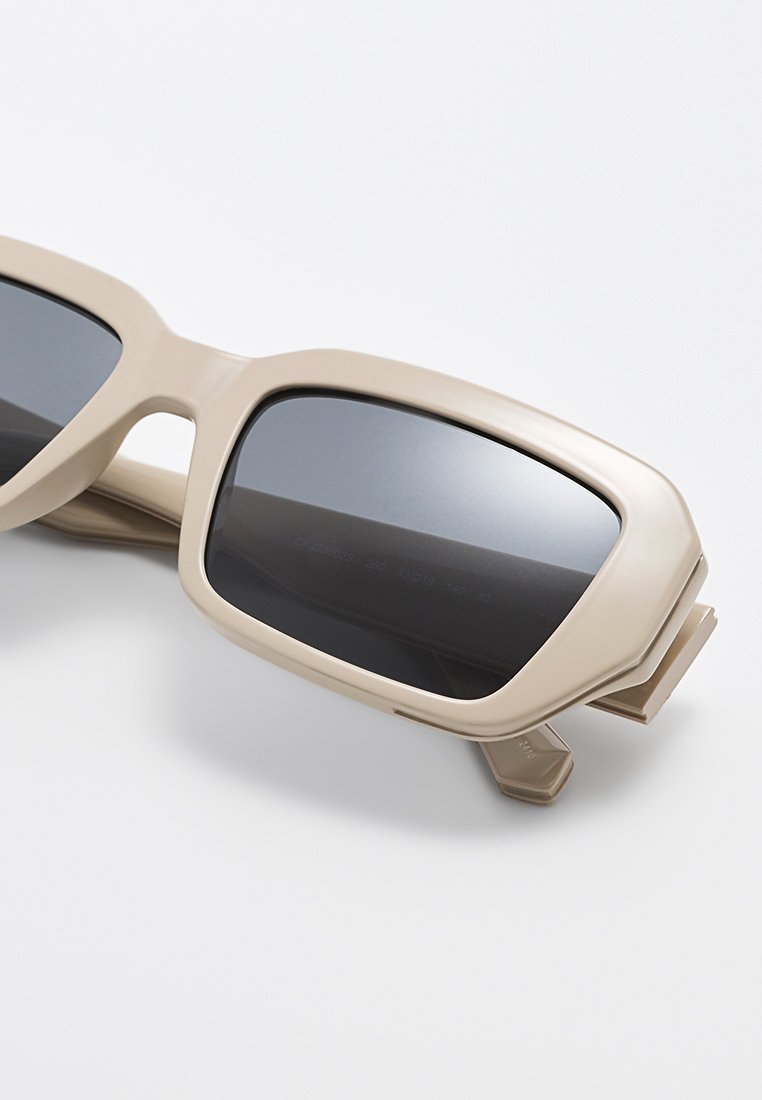 Beżowe, przerośnięte okulary przeciwsłoneczne z kwadratowymi oprawkami, ciemno przyciemnionymi soczewkami oraz matowym wykończeniem. Charakteryzują się grubym, aerodynamicznym designem i brakiem widocznych oznakowań.