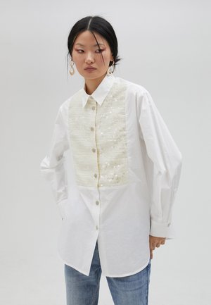 White shirt with a textured cream panel, featuring sequins and buttons down the front. Long sleeves and a classic collar design.
