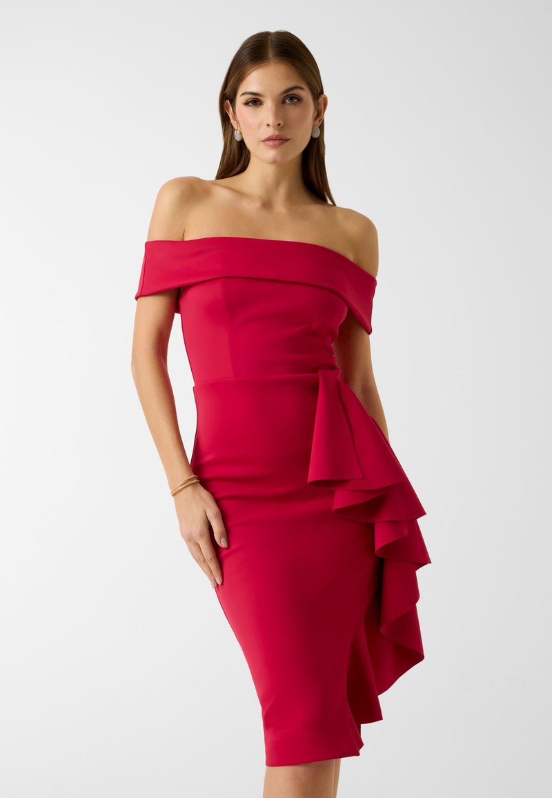 Femme portant une robe rouge ajustée à épaules dénudées avec un détail à volants superposés sur le côté droit, debout devant un fond uni.