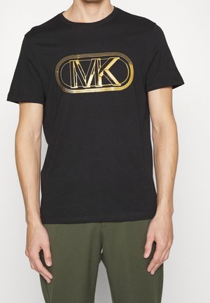 Homme portant un T-shirt noir avec un logo "MK" en or métallique et un pantalon vert olive, debout devant un fond uni.