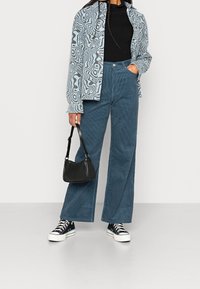 Blått plyschbyxor med vida ben och en texturerad yta, i kombination med en mönstrad ljusblå skjorta och svart topp, kompletterat med svarta sneakers och väska.