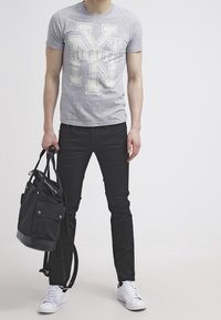 Grå kortärmad t-shirt med vit logotyp, svarta slim-fit jeans, vita sneakers och en svart tygväska med flera fickor.