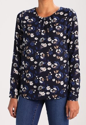 Granatowa bluzka z długim rękawem w biało-niebieski kwiatowy wzór, noszona z niebieskimi dżinsami, widoczna od szyi do bioder.