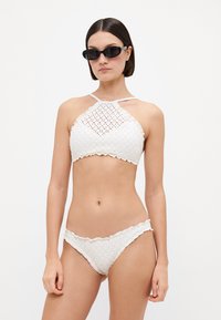 Biely háčkovaný bikiny set s halter krkom a vykrojenými okrajmi, doplnený o zodpovedajúce spodky. Hladká textúra, zložené vzorové detaily.