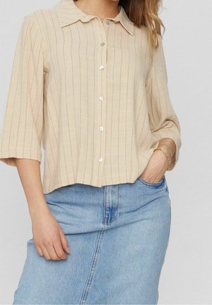 Vrouw in beige, verticaal gestreept overhemd met knopen en driekwart mouwen, in een lichtblauwe spijkerrok gestopt, met gouden kettingarmbanden aan haar rechterpols.