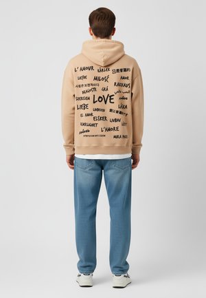 Felpa con cappuccio beige con testo nero in più lingue sulla schiena. Abbinata a jeans azzurri chiari e sneakers bianche.