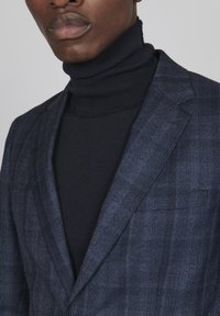 Blå rutig kavaj över en svart rullkrage. Kavajen har en texturerad tyg med påfallande mönsterlinjer och strukturerade slag.