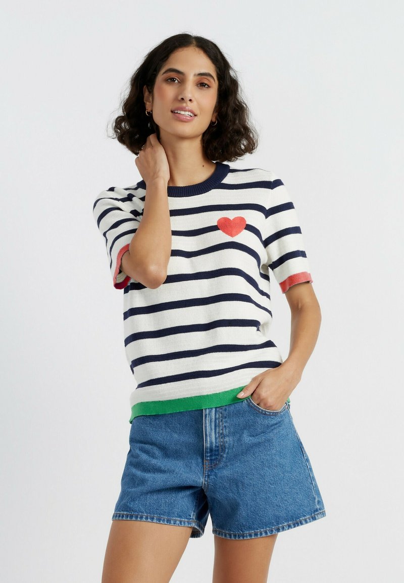 Pull en tricot à manches courtes avec des rayures bleu marine et blanches, un détail cœur rouge et un ourlet vert. Associé à un short en denim bleu.