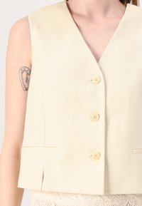Beige sleeveless vest with a deep V-neck, featuring four yellow buttons, a textured fabric, and side pockets at the hem.