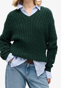 Green cable-knit sweater with a V-neck, layered over a light blue button-up shirt, pairing with light blue jeans and a brown belt.