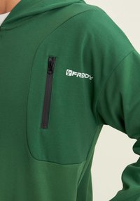 Sudadera verde hecha de una tela suave, con un bolsillo con cremallera en el pecho, un acento de logo en el lado y un diseño ajustado.
