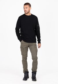Felpa nera a maniche lunghe con texture a coste, scollatura rotonda e tasca con zip sulla manica sinistra; abbinata a pantaloni cargo verdi e stivali neri.