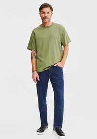 Oliwkowa bawełniana koszulka z krótkim rękawem, połączona z ciemnoniebieskimi dżinsami i czarnymi sneakersami. Prosty, luźny krój, minimalne detale.