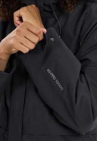 Giacca nera impermeabile con texture liscia, polsini con zip e bottone a pressione, e logo "W-PRO 10000" stampato sulla manica.