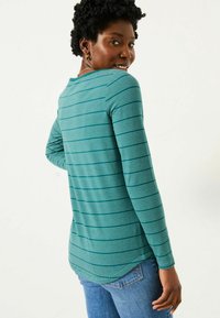 Frau mit kurzem lockigem Haar, die ein gestreiftes, langärmeliges Shirt in Türkis und Blue Jeans trägt. Sie schaut über die Schulter und hat ein leichtes Lächeln.