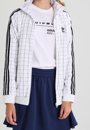 Personne portant une veste blanche Adidas avec des rayures noires et un motif quadrillé, un t-shirt blanc Adidas et une jupe bleu marine.