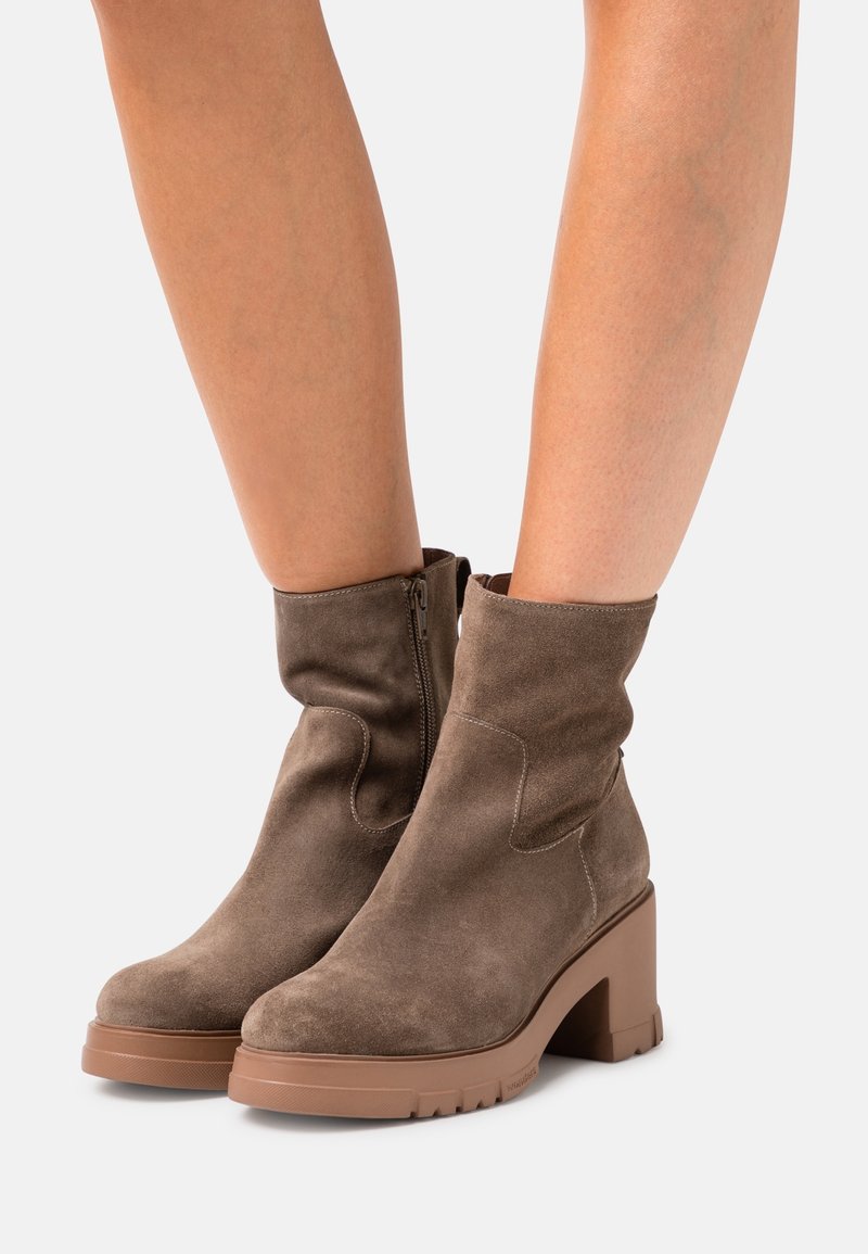 Bottines en daim marron avec talons carrés et fermetures éclair latérales, portées sur des jambes nues sur un fond uni.