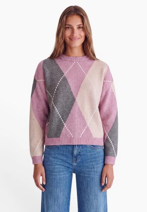 Giovane donna che indossa un maglione con motivo argyle rosa, grigio e beige e jeans blu, in piedi e sorridente su uno sfondo bianco.
