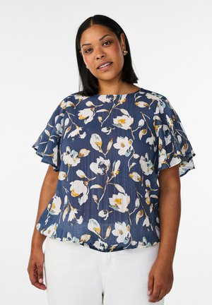 Kvinde iført en mørkeblå bluse med hvidt og gult blomsterprint og flagrende ærmer, kombineret med hvide bukser, står foran en ensfarvet baggrund.