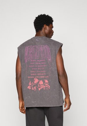 Mand iført en ærmeløs, falmet sort T-shirt med pink "WORLD TOUR" tekst, datoer og kraniumgrafik på ryggen, vendt væk fra kameraet.