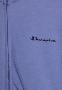 Gaiši purpursarkans kapuci džemperis ar rāvējslēdzēju, izgatavots no mīkstas auduma, ar melnu "Champion" logotipu kreisajā krūtīs un teksturētām ribu detaļām.