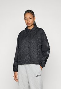 Svart mönstrad jacka med krage och knappstängning. Har elastiska ärmslut och sidor med texturerad blommönsterdesign. Matchad med gråa sweatpants.