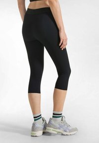 Leggings capri neri con una vita ampia e liscia, realizzati in tessuto elasticizzato. Indossati con sneakers grigie e lavanda e calzini a righe.