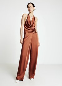 Braunes Satin-Halfter-Top mit drapiertem Ausschnitt, kombiniert mit hochtaillierter brauner Hose. Weiße spitze Absatzschuhe und ein dekorativer Knopfakzent.