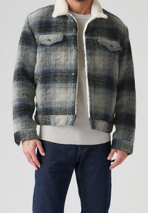 Mężczyzna ubrany w niebiesko-szarą kurtkę w kratę z białą podszewką sherpa, jasnoszary teksturowany sweter i ciemnoniebieskie dżinsy.