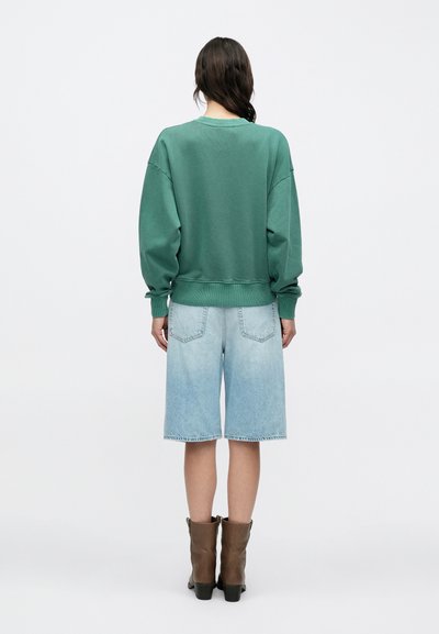 Γυναίκα με την πλάτη γυρισμένη, φοράει πράσινο φαρδύ φούτερ, ανοιχτόχρωμα σορτς μέχρι το γόνατο από τζιν και καφέ μπότες μέχρι τη μέση της γάμπας σε λευκό φόντο.