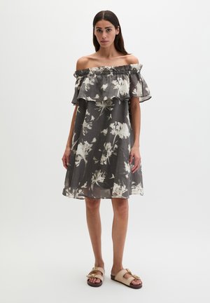 Femme portant une robe fleurie gris foncé avec épaules dénudées et des sandales beiges, debout devant un fond blanc uni.