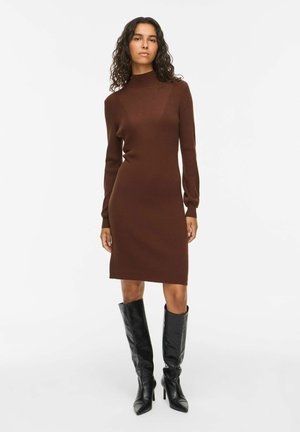 Femme aux cheveux bouclés portant une robe en maille marron cintrée à manches longues et des bottes noires à talons montantes jusqu'aux genoux, debout sur un fond blanc.