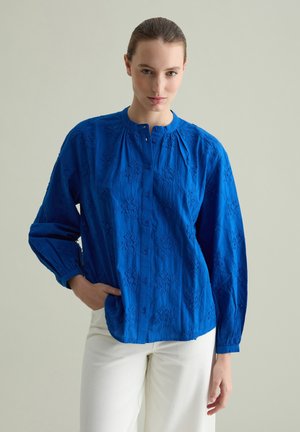 Camicia blu con bottoni, decorata con un motivo floreale testurizzato, maniche lunghe e scollo arricciato. Abbinata a pantaloni bianchi.