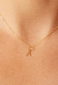 Goldene Halskette mit einem einzigen polierten Buchstaben "A" als Anhänger, der auf einer glatten Hautoberfläche liegt. Zartes Design mit feiner Struktur.