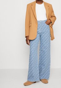 Blazer marrone chiaro sopra una maglia bianca, abbinato a pantaloni ampi blu con motivi astratti, completato da sandali pelosi color cuoio.