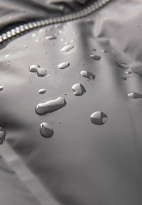 Vodotesný šedý materiál s viditeľnými kvapkami. Hladký povrch s jemným leskom. Detaily so zipsom blízko hornej hranice obrázka.