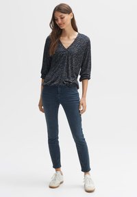 Marinebluse mit V-Ausschnitt, Drei-Viertel-Ärmeln und einem strukturierten Muster, kombiniert mit dunkelblauen Jeans und weißen Turnschuhen.