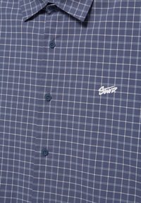 Modra karo srajca z gumbi, z belo mrežasto vzorcem in belim vezenim logotipom "Sport" na prsih.