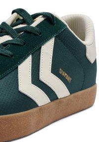 Zelen športni čevelj z teksturirano tkanino, belimi cik-cak poudarki in gumijastim podplatom. Ima zavezovanje z vezalkami in napis "DIAMANT" v zlati barvi.