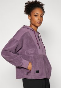 Haut en molleton violet avec capuche et fermeture éclair sur le devant. Présente un tissu texturé et une poche kangourou. Accentué par des cordons de serrage noirs et une étiquette.
