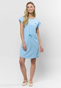 Šviesiai mėlyna trumpų rankovių suknelė, pagaminta iš minkštos medžiagos, su raišteliu juosmenyje ir mažuoju logotipu šalia iškirptės.
