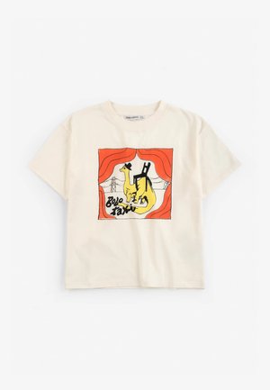 Camiseta crema con un gráfico de un dinosaurio amarillo que lleva un sombrero negro, de pie bajo una cortina roja con una silla negra en su espalda.