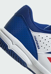 Chaussure de sport bleue et blanche avec texture perforée, semelle en caoutchouc et accents rouges. Comprend un col rembourré et un matériau en maille pour la respirabilité.