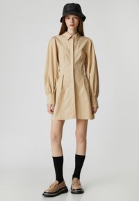 Beiges Hemdkleid mit langen Puffärmeln, Knopfleiste und Seitentaschen. Getragen mit schwarzen Kniestrümpfen und braunen Slippern. Schwarzer Bucket Hat.